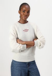 Sweatshirt gris avec des accents crème et un col bordeaux. Présente "Tommy HILFIGER" brodé en rouge. Coupe décontractée, manches texturées.