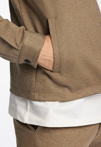 Giacca in tessuto marrone con texture, dotata di zip, tasche laterali e uno strato bianco sottostante, caratterizzata da una finitura a motivo sottile.