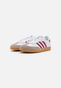White sneakers with red stripes, grey suede toe cap, and tan rubber sole. Features a lace-up design and blue accent logo on tongue.