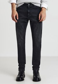 Uomo che indossa jeans neri sbiaditi, un maglione grigio con le maniche arrotolate e stivali in pelle nera, in piedi su un pavimento chiaro.