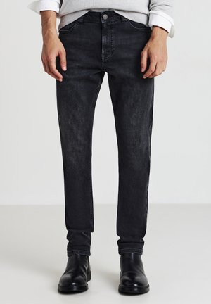 Homme portant un jean noir délavé, un pull gris à manches retroussées et des bottes en cuir noires, debout sur un sol clair.