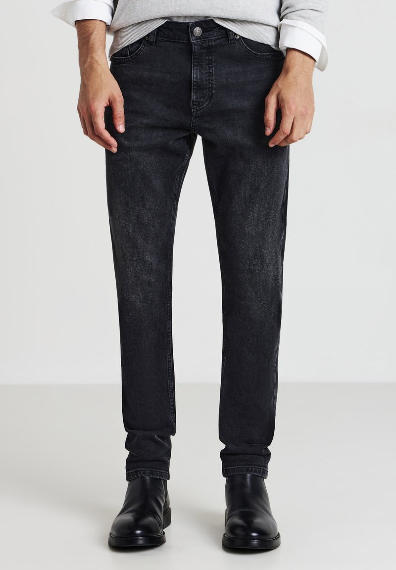 Uomo che indossa jeans neri sbiaditi, un maglione grigio con le maniche arrotolate e stivali in pelle nera, in piedi su un pavimento chiaro.