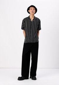 Cămașă scurtă cu mâneci, cu dungi alb-negru, design cu nasturi, având o croială lejeră, purtată cu pantaloni negri largi și pantofi negri masivi.