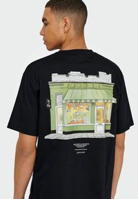 Sort t-shirt med et farverigt grafik af en butik med navnet "FRANKS" med en grøn facade, stribet markise og vinduesudstillinger.