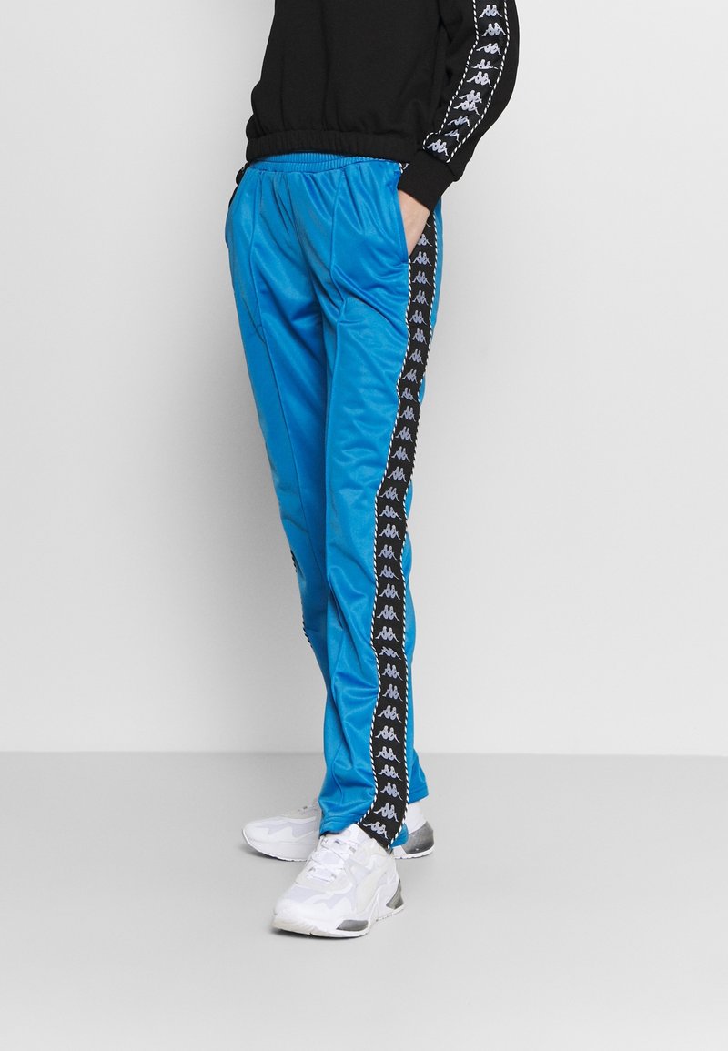 Blå sportbyxor med en glänsande textur, elastisk midjeband, svarta sidostripes med ett logomönster, tillsammans med vita sneakers.