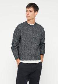 Σκούρο γκρι πλεκτό πουλόβερ με υφή, στρογγυλή λαιμόκοψη και ριχτές μανσέτες. Φορεμένο πάνω από λευκό πουκάμισο, συνδυασμένο με σκούρες παντελόνες.