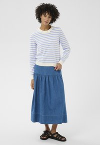 Femme aux cheveux bouclés portant un pull rayé blanc et bleu clair, une longue jupe en jean et des sandales noires, debout devant un fond uni.