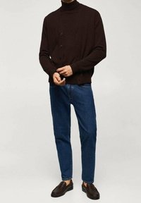 Donkerbruine, gebreide cardigan met een col, sluiting met knopen, gecombineerd met een blauwe spijkerbroek en donkere instapschoenen.