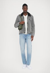 Veste en velours côtelé grise avec col noir, T-shirt blanc, jeans bleu clair et baskets blanches ; tenant un sac bandoulière vert olive.