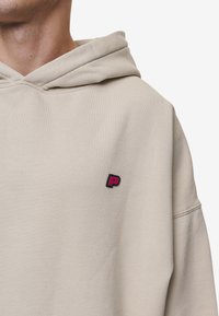Sweatshirt bege com cordão e um pequeno logótipo preto e rosa no peito. Apresenta um tecido suave e texturizado, com um corte solto.