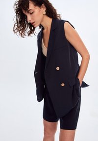 Námořnický bezrukávový blazer s dřevěnými knoflíky, dvěma předními kapsami a volnějším střihem, doplněný o odpovídající šité kraťasy.