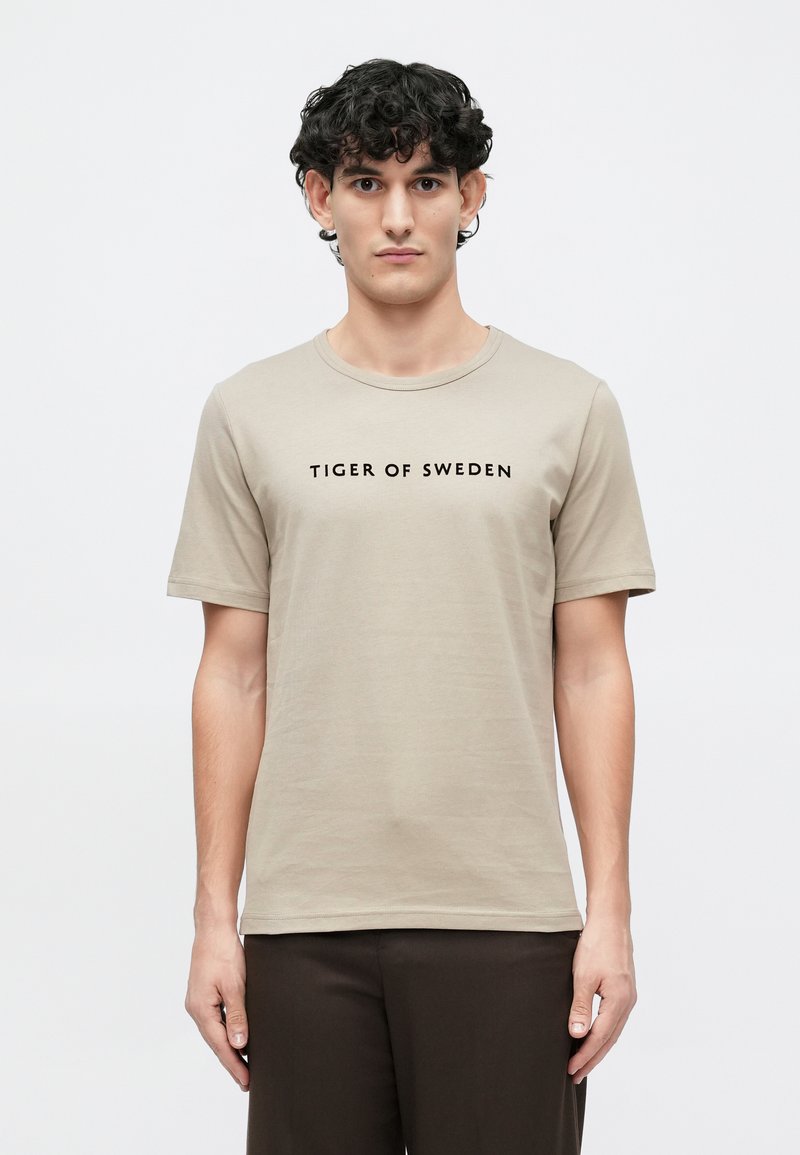 Béžové tričko s krátkym rukávom vyrobené z jemnej látky, s okrúhlym výstrihom, s potlačeným čiernym logom "TIGER OF SWEDEN" na hrudi.