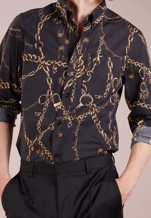 Schwarzes Hemd zum Knöpfen mit Muster aus goldener Kette und Sicherheitsnadel, hochgekrempelte Ärmel, getragen mit schwarzer Hose und Händen in den Taschen.