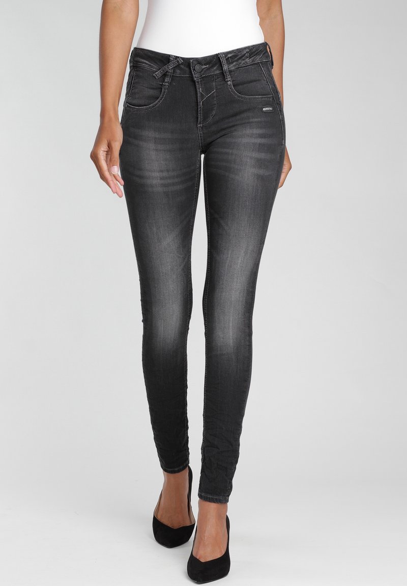 Vaqueros skinny negros con un acabado desgastado, que cuentan con cierre de botón, bolsillos frontales y un ajuste elegante, combinados con tacones altos negros.
