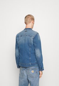 Man som står med ryggen mot kameran, iklädd en bleknad blå denimjacka och matchande jeans med synliga märkeslappar på bakfickorna och kragen.