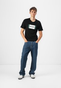 Svart bomull T-skjorte med en hvit og grønn logo-patch, kombinert med lyseblå løstsittende denimjeans og hvite kondiser med svarte detaljer.