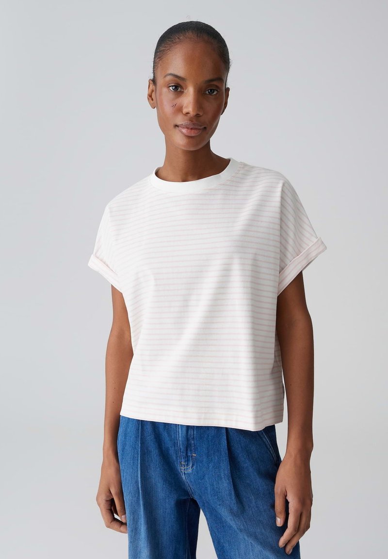 Femme portant une chemise blanche à manches courtes avec des rayures rose clair et un jean bleu, debout devant un fond gris clair uni.