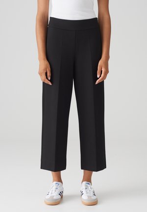 Person iført sorte vidde ben, cropped bukser, en hvid ærmeløs top og hvide sneakers med sorte striber, stående mod en ensfarvet baggrund.