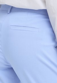 Pantalon bleu clair ajusté avec une poche passepoilée à l'arrière et des passants pour ceinture, porté avec une chemise blanche rentrée.