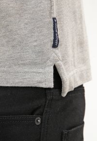 Šedé bavlněné tričko s bočním rozparkem, na němž je modrá látková značka s nápisem „Armani Jeans“. V kombinaci s černými džíny a detaily na knoflících.