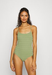 Femme aux cheveux bouclés et foncés portant un maillot de bain une pièce texturé de couleur vert clair avec des fines bretelles et un lien sur le côté, se tenant devant un fond uni.