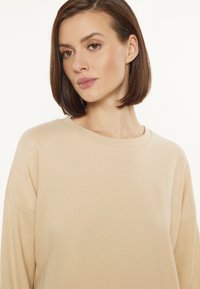 Beige színű pulóver, laza fazonnal, kerek nyakkal és ejtett vállakkal. Puha szövet textura; nincs látható minta vagy fémalkatrész.