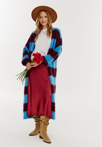 Pasiasty kardigan w niebieskim i burgundowym kolorze, z długimi rękawami, noszony na białym T-shircie i bordowej spódnicy. Brązowy kapelusz z szerokim rondem i beżowe buty do kolan.