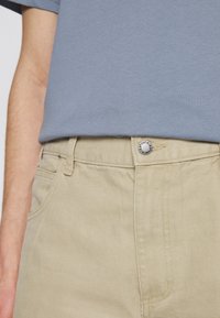 Beige pants with a textured finish and a metal button at the waistband. The outfit features a light blue shirt.