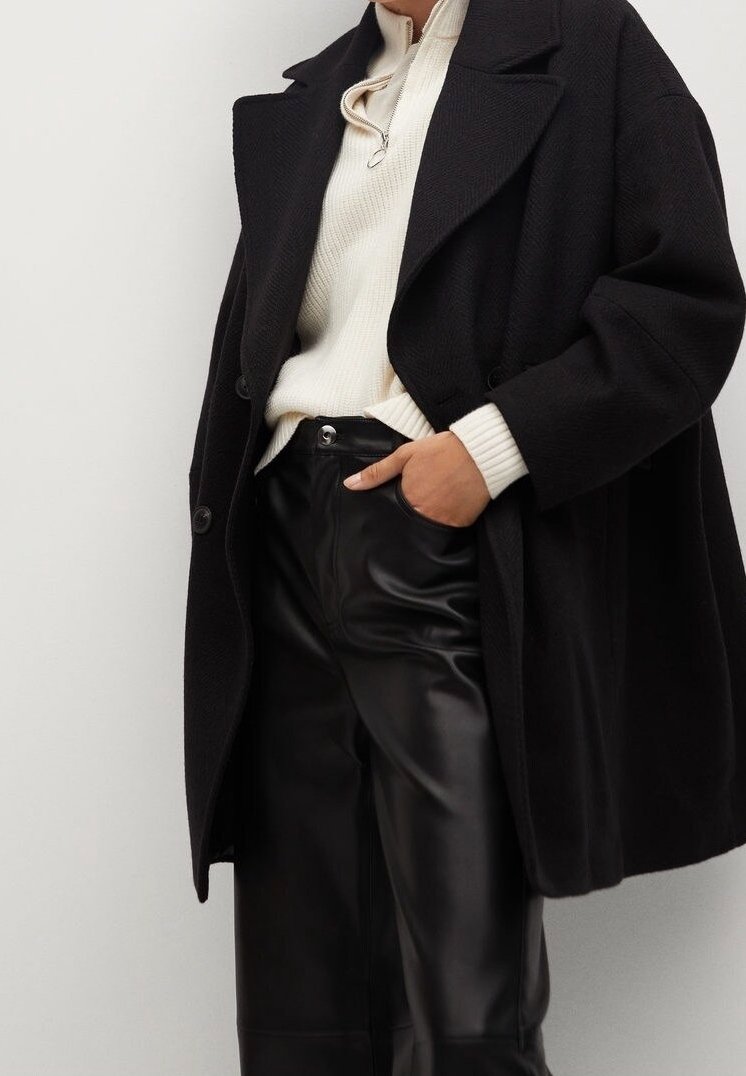 Personne portant un manteau noir oversize, un pull en laine crème avec fermeture éclair, et un pantalon en cuir noir, main posée négligemment dans la poche sur un fond uni.