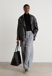 Svart skinn bikerjakke, tettsittende svart genser med høy hals, lys grå plissert maxiskjørt, svart tote bag, grå blokkhæler, nøytral bakgrunn.