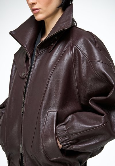 Detailný záber na osobu nosiacu tmavohnedú nadrozmernú koženú bundu s vysokým golierom, zipsom vpredu a elastickými manžetami, ruky v kapsách.