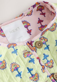 Veste en tissu matelassé vert avec motif floral orange et rose, doublure rose, boutons en bois et étiquette portant la mention "NEXT".