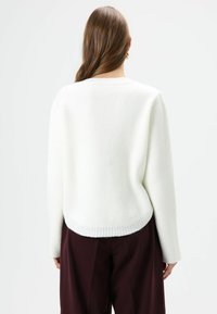Pull tricoté blanc avec un col rond, des épaules tombantes et un ourlet côtelé. La texture semble douce, avec une coupe décontractée et un design minimaliste.