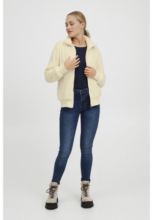 Veste zippée crème texturée avec un col haut, associée à un haut bleu marine et un jean bleu. Le mannequin porte des bottes de randonnée beiges.