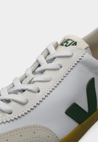 Λευκό αθλητικό παπούτσι με πράσινες λεπτομέρειες και κορδόνια. Διαθέτει υφασμάτινη επένδυση με υφή, αντίθετο γκρι πάνελ και ορατή ραφή σε ζιγκ ζαγκ κοντά στη σόλα.