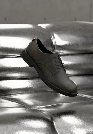 Σκούρο γκρι δερμάτινο παπούτσι με κορδόνια που αιωρείται πάνω από μια λαμπερή ασημί μεταλλική επιφάνεια με κουμπιά, με μαλακό φωτισμό που αναδεικνύει τις υφές.