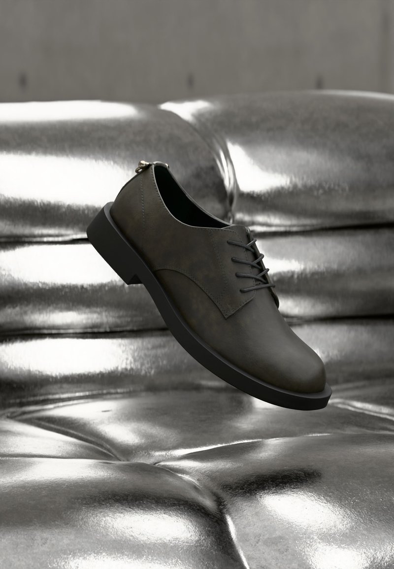 Σκούρο γκρι δερμάτινο παπούτσι με κορδόνια που αιωρείται πάνω από μια λαμπερή ασημί μεταλλική επιφάνεια με κουμπιά, με μαλακό φωτισμό που αναδεικνύει τις υφές.