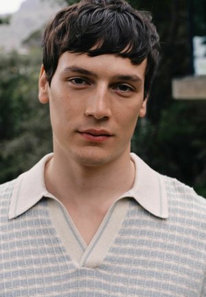Young man with short dark hair wearing a light-colored, textured knit shirt with a collar, standing outdoors with blurred greenery behind him.