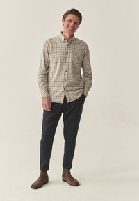 Cămașă în carouri bej și maro, combinată cu pantaloni bleumarin și cizme scurte maronii. Cămașa are un guler clasic și nasturi pe față.