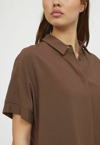 Chemise marron à manches courtes avec un col boutonné, présentant une texture lisse et une coupe classique, mise en valeur par des détails de couture subtils.