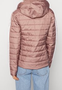 Jasnoróżowa, pikowana kurtka puchowa z kapturem, z białymi paskami po bokach i gładką fakturą, noszona na niebieskich dżinsach.