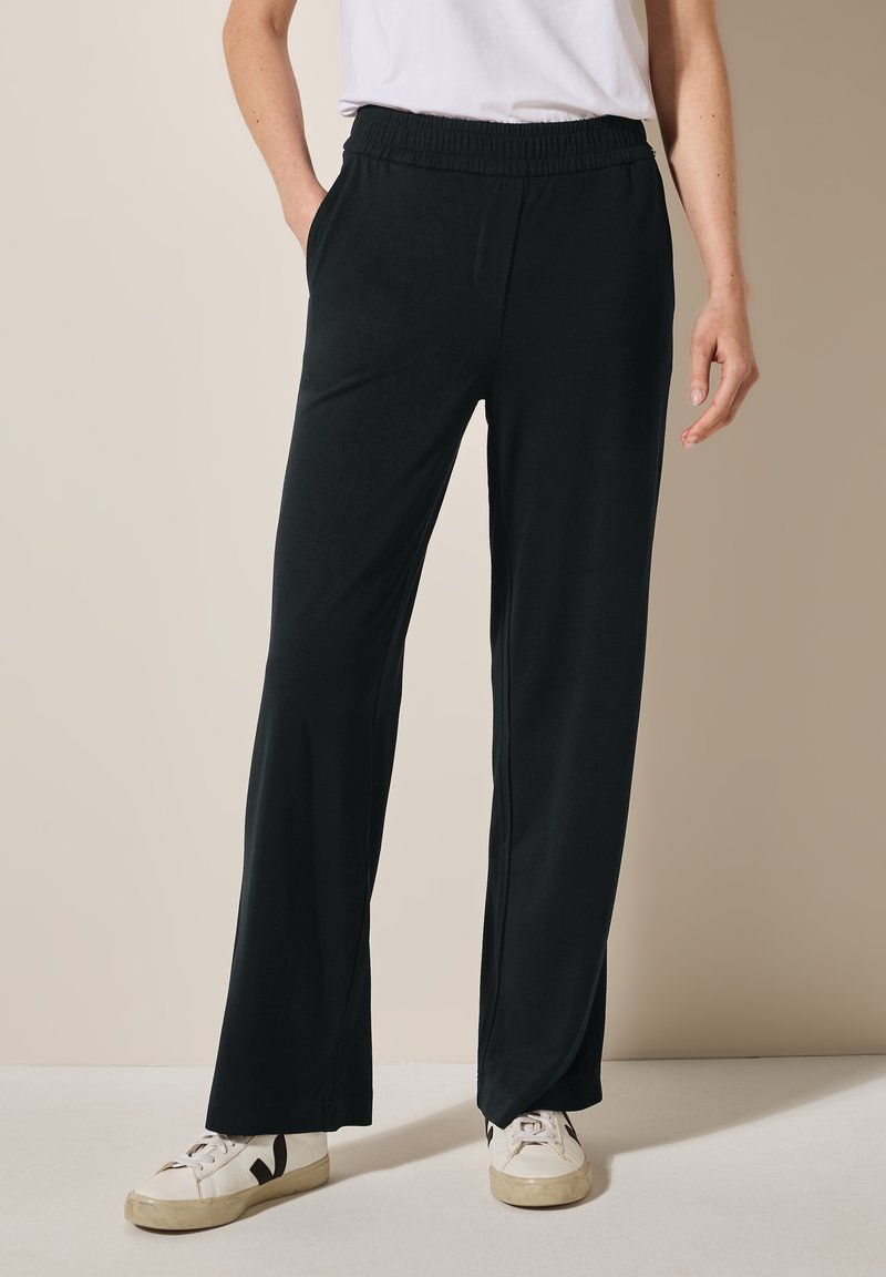 Zwarte, lichte, wijde broek met een elastische tailleband en zijzakken, gecombineerd met witte sneakers. Glad textuur en minimalistisch ontwerp.