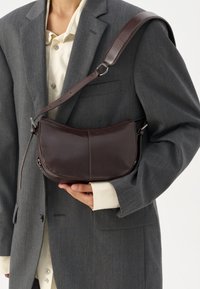 Dark brown leather shoulder bag with a curved shape, thin strap, and subtle stitching details. Worn over a gray blazer and cream shirt.