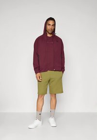 Bordo jopica z zadrgo, z gorom in stranskimi žepi, v kombinaciji z olivno zelenimi kratkimi hlačami. Pogled zaključujejo beli športni copati in sive nogavice z logotipom.