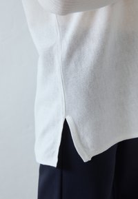 Valkoinen neulepaita pyöreällä helmalla, jossa on sivuhalkiot. Kangas on pehmeää hienorakenteista, ja se on yhdistetty tummansinisiin housuihin.