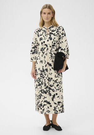 Mujer con un vestido largo color crema con estampado floral negro, sosteniendo una prenda negra doblada, de pie frente a un fondo blanco liso.