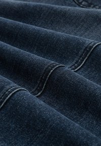 Tela de mezclilla azul oscuro con textura suave, que presenta costuras visibles y pliegues. El material muestra ligeras variaciones de color y un brillo suave.