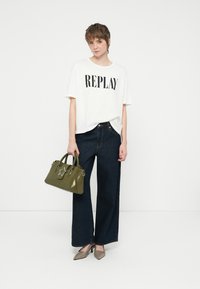 Camiseta branca de mangas curtas com "REPLAY" em impressão preta, combinada com jeans de perna larga em denim escuro, bolsa verde com detalhe de estrela e saltos bejes.