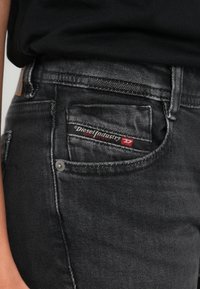 Personne portant un jean noir avec une étiquette Diesel Industry visible sur la petite poche avant, assorti à une chemise noire.