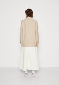 Beige σακάκι με κοπή, με έναν αεραγωγό, φορεμένο πάνω από μια μακριά λευκή φούστα. Εμφανίζει λεπτομέρειες κουμπιών στα μανίκια και λευκά αθλητικά παπούτσια.